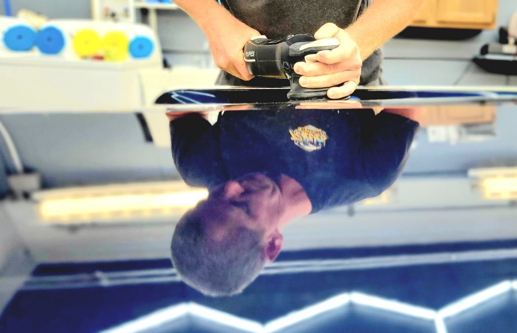 A technician polishing a black vehicle surface with a dual-action polisher, reflecting his Retro Detailing shirt and workshop lighting.