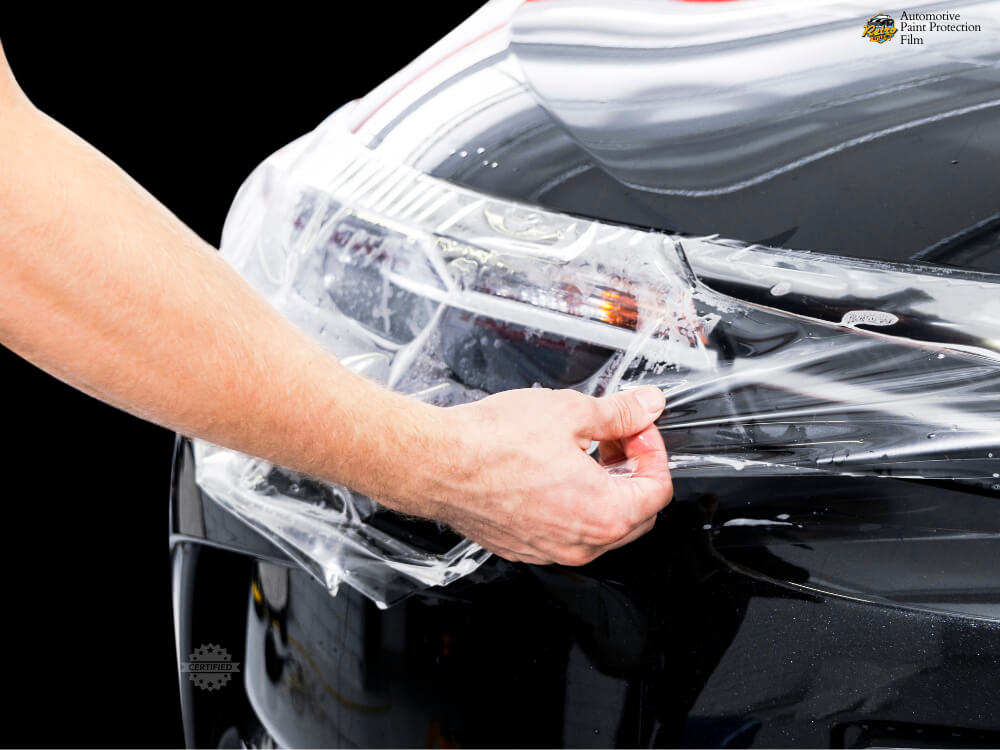 Hand applying clear paint protection film (PPF) to the front headlight of a black vehicle.